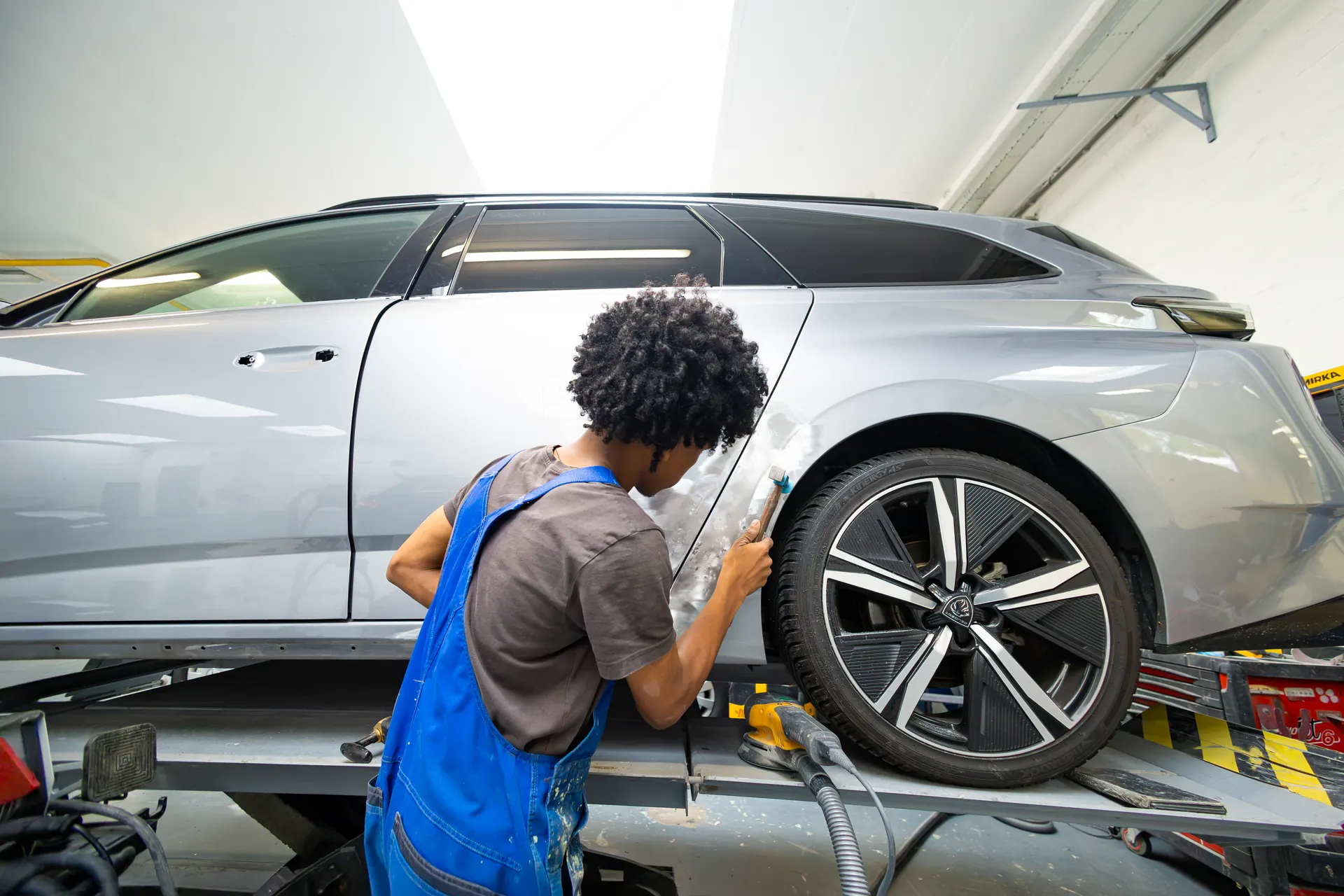 Een monteur in een blauwe overall werkt aan een deuk in het achterportier van een grijze auto.