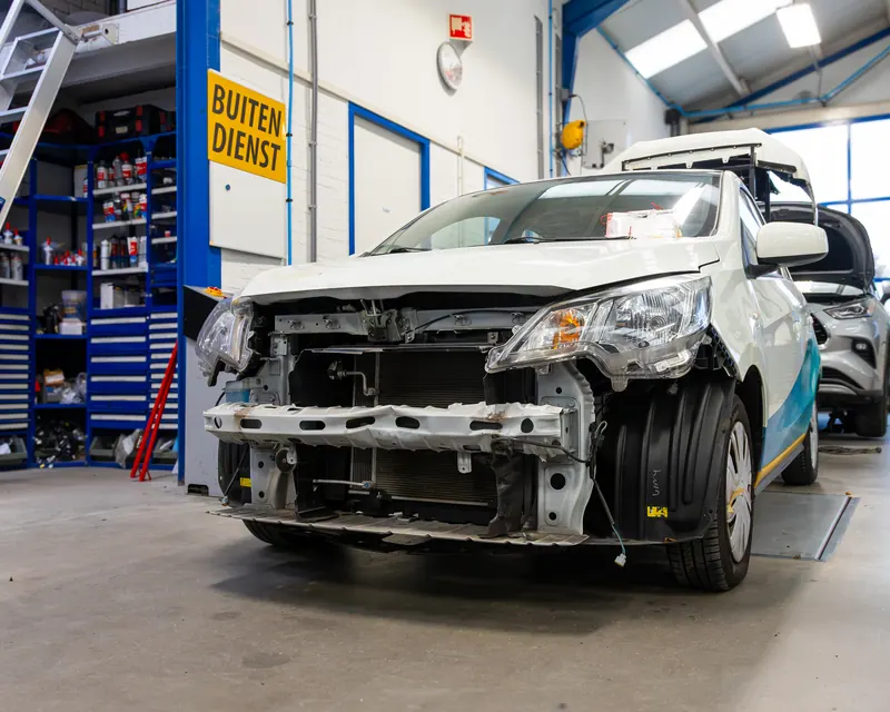 Een witte auto met een beschadigde voorkant in de werkplaats, klaar om op te nemen.
